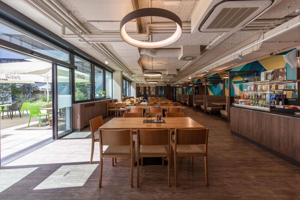 Helle Cafeteria mit Holztischen, modernen Stühlen und Zugang zu einer Außenterrasse.