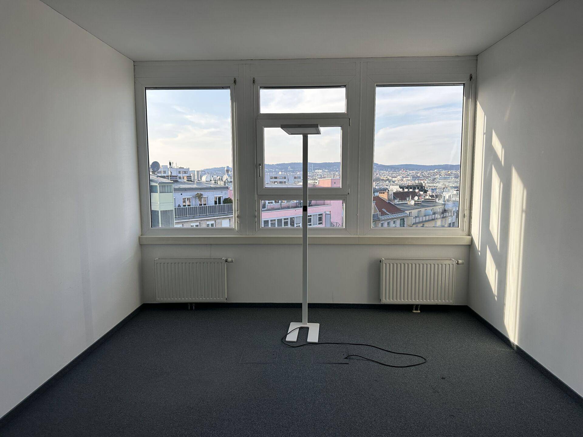 Leeres Büro mit Teppichboden, großen Fenstern und Heizkörpern, bietet einen weiten Stadtblick.