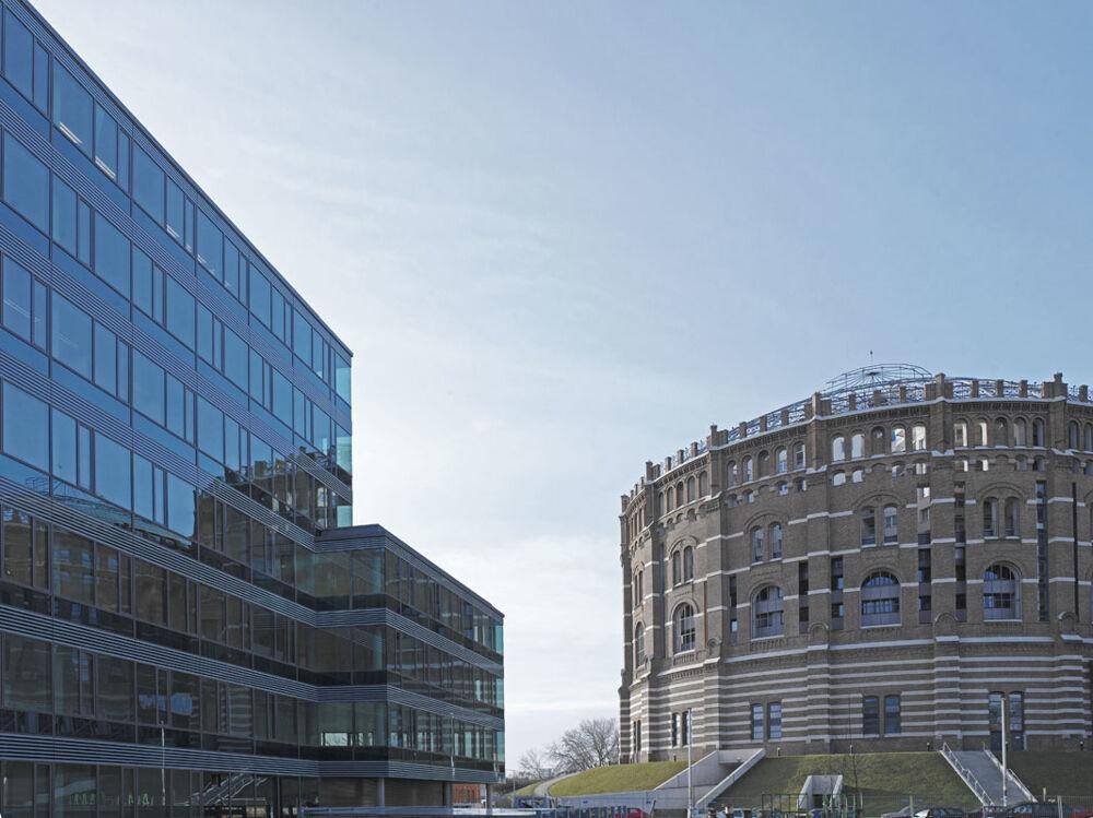 Moderne Glasfassade eines Bürogebäudes neben einem historischen Gasometer unter blauem Himmel.