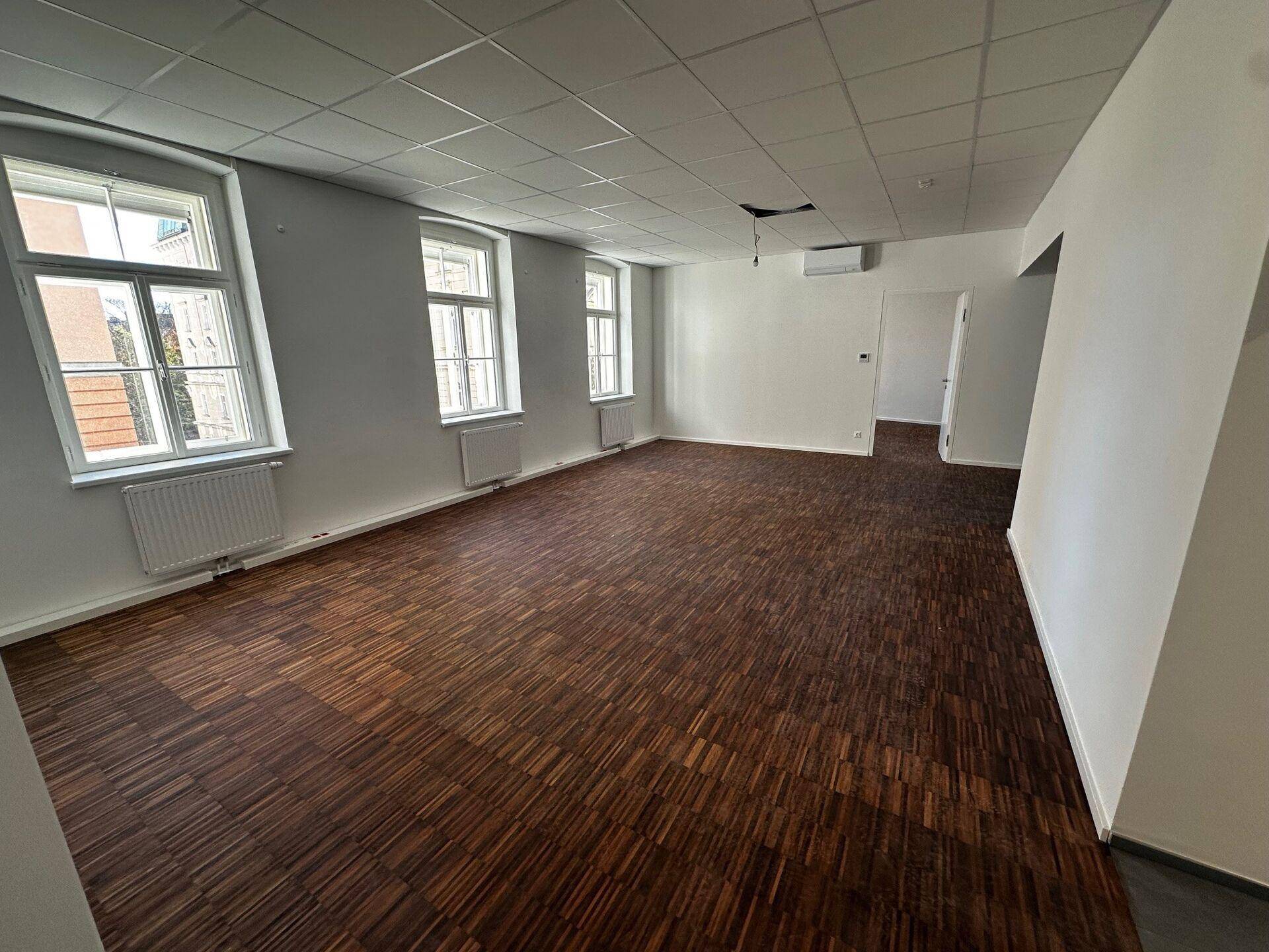 Großes, leeres Büro mit dunklem Holzboden, weißen Wänden und abgehängter Decke.