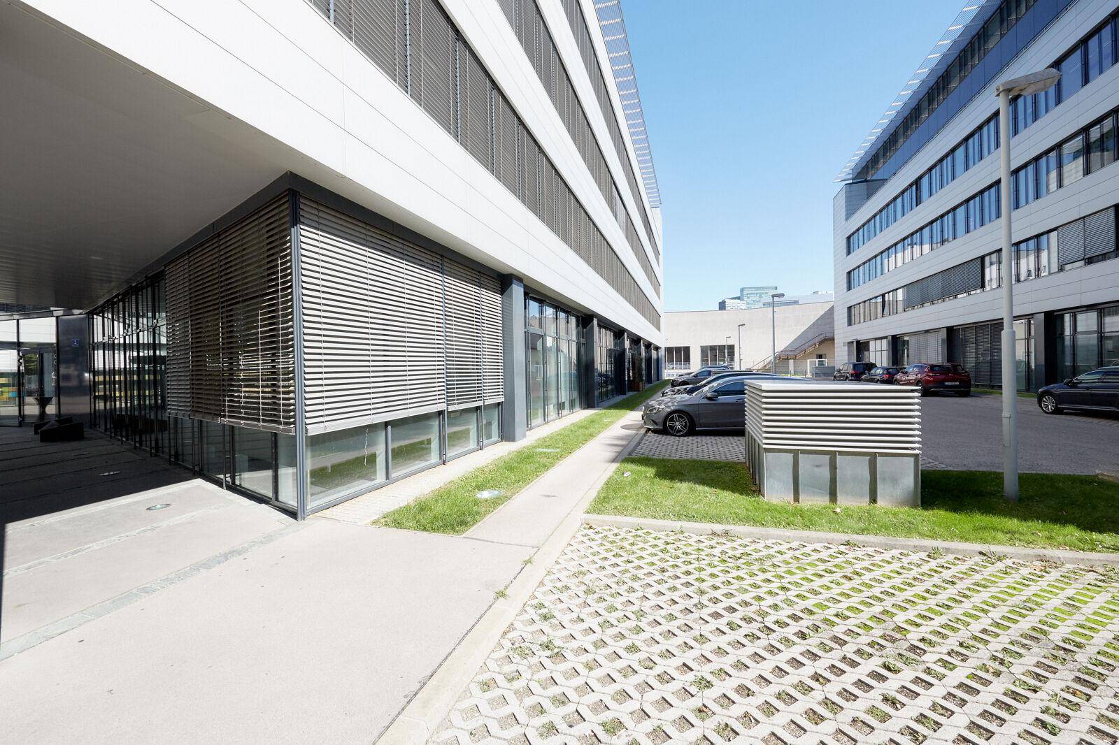 Außenbereich eines Bürogebäudes mit Parkplätzen, Rasenflächen und Jalousien an den Fenstern.