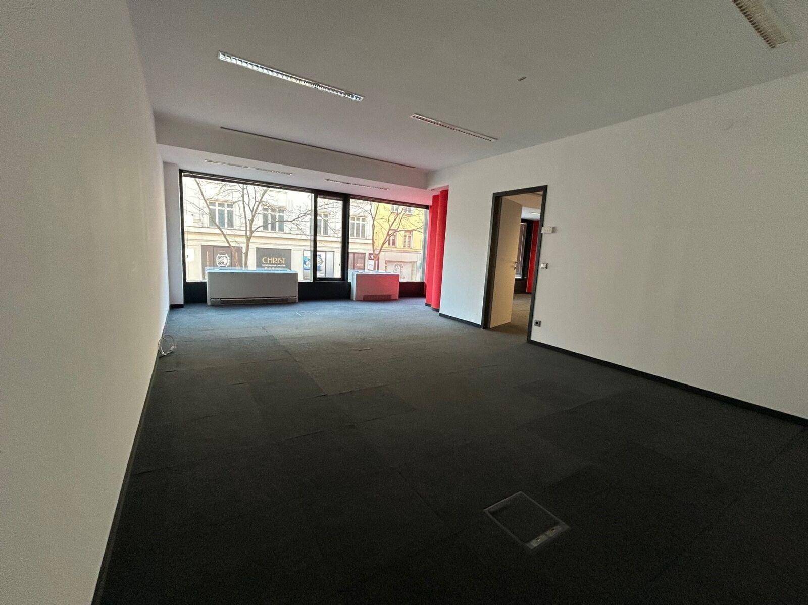 Heller Bürobereich mit Teppichfliesen, großen Fenstern und Zugang zu weiteren Räumen.