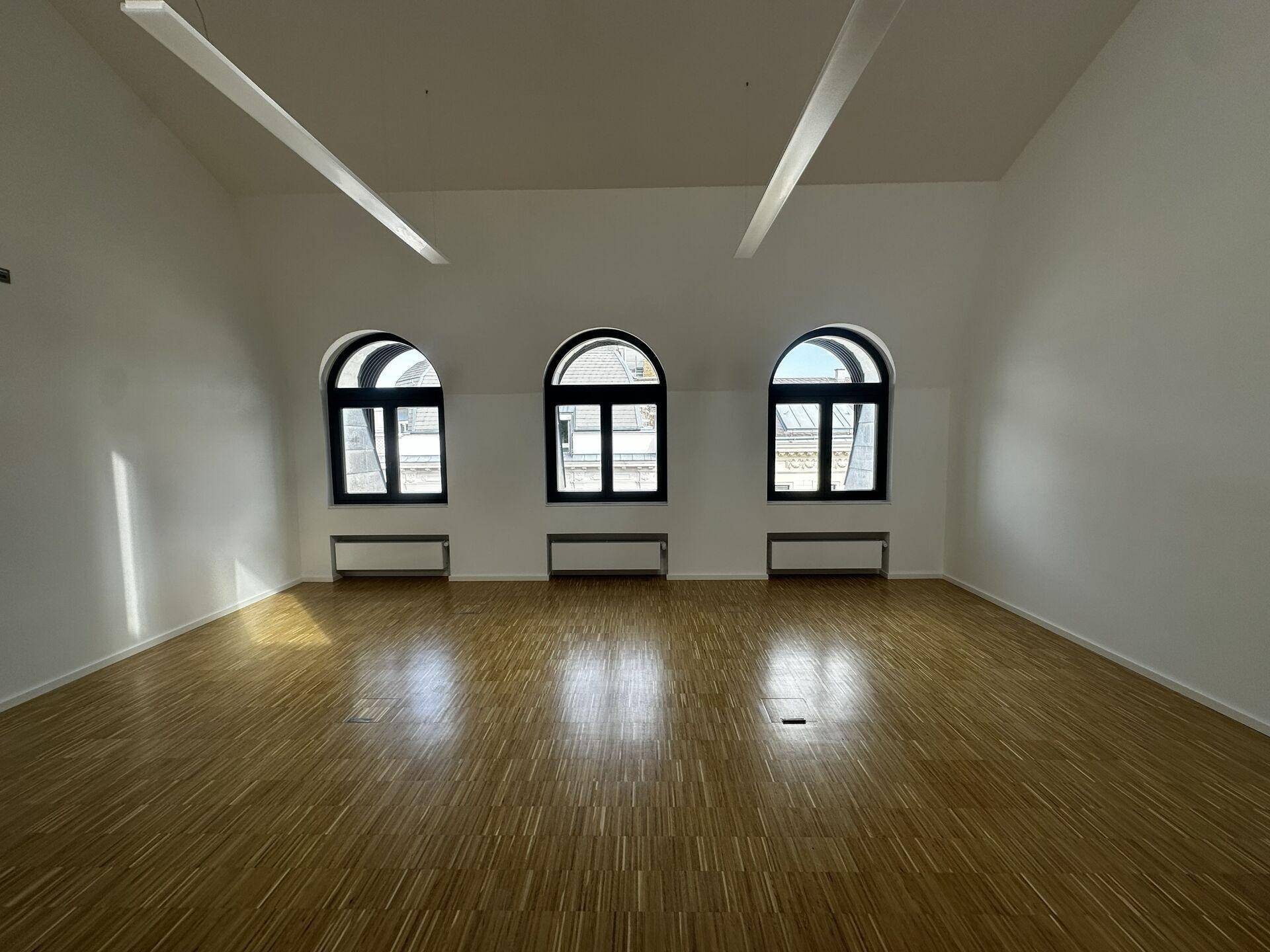 Heller Büroraum mit Holzboden, hohen Decken und drei eleganten Rundbogenfenstern.