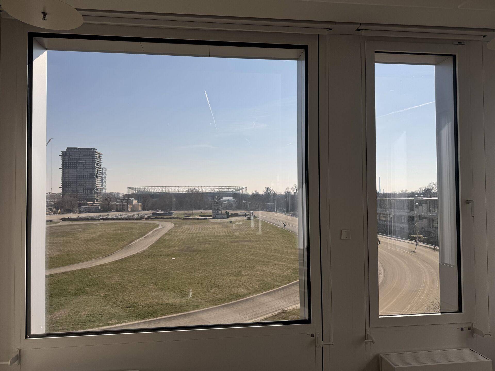 Fensterblick aus einem Büro auf eine weite Grünfläche und städtische Gebäude in der Ferne unter klarem Himmel.