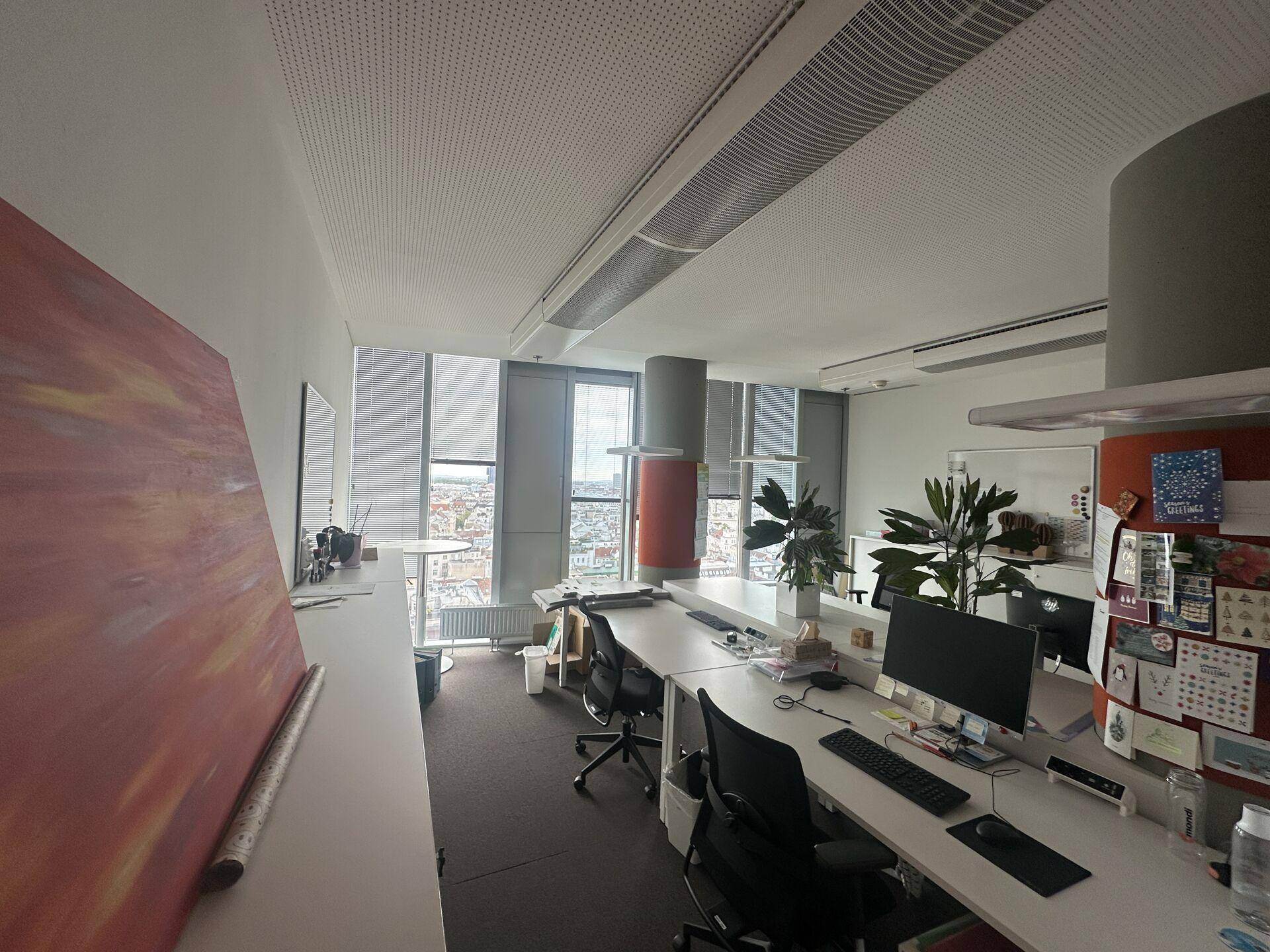 Offenes Büro mit mehreren Arbeitsplätzen, modernen Möbeln und großen Fenstern, die einen weiten Blick über die Stadt bieten.