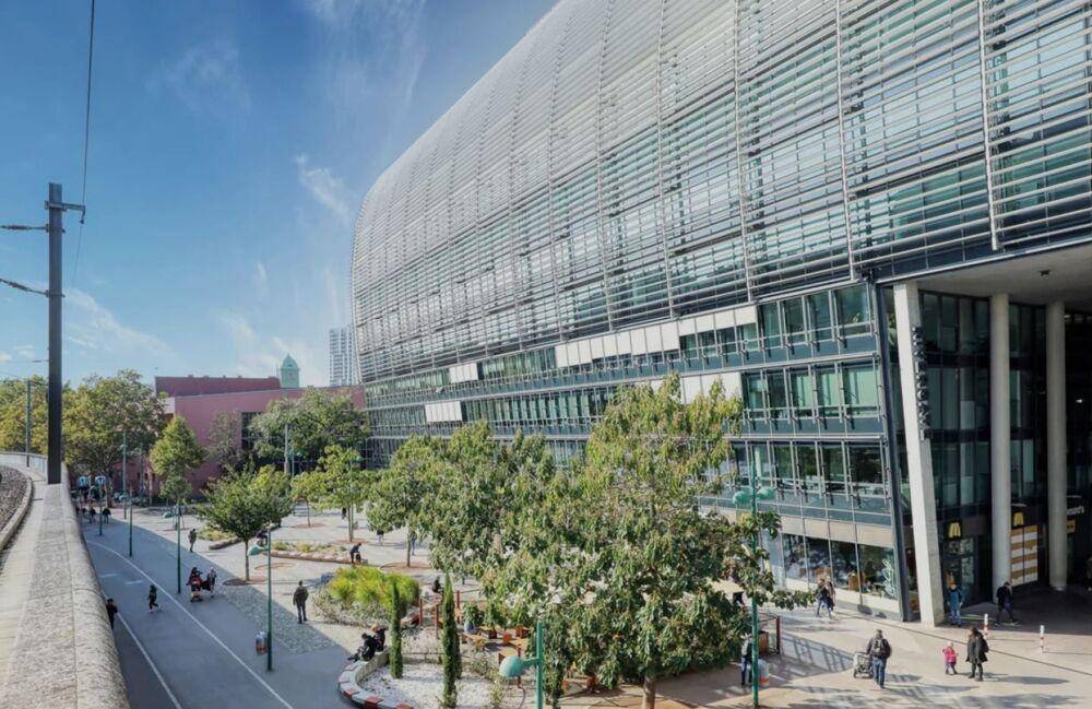 Ansicht eines modernen Bürokomplexes mit breitem Gehweg, Grünflächen und Passanten an einem sonnigen Tag.