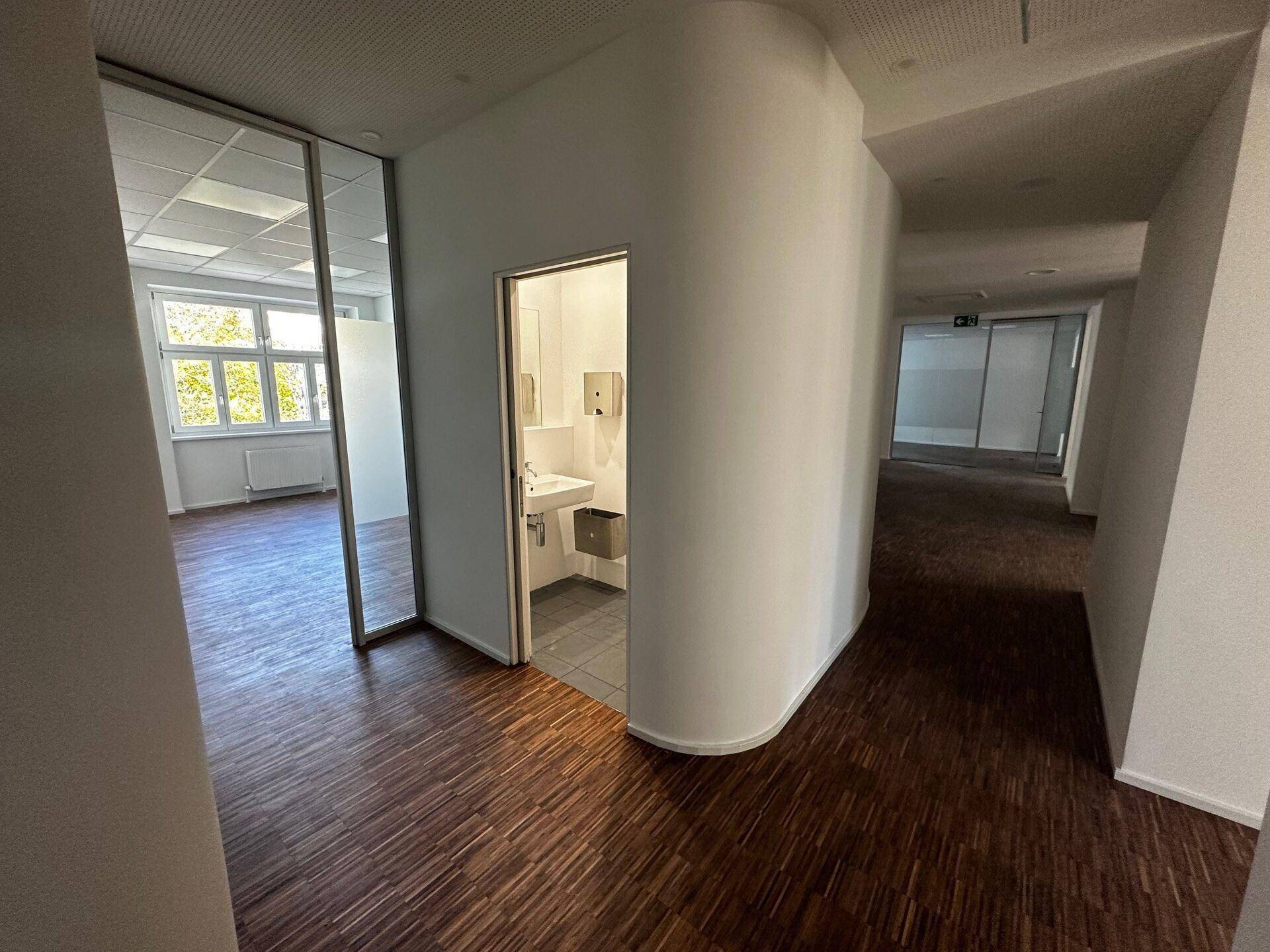 Heller Flur in einem Bürogebäude mit Holzboden, Glastüren und Zugang zu einem kleinen Bad.