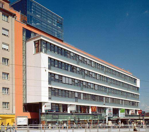 Ansicht eines modernen Bürogebäudes mit großen Fenstern und orangefarbenen Akzenten unter blauem Himmel.