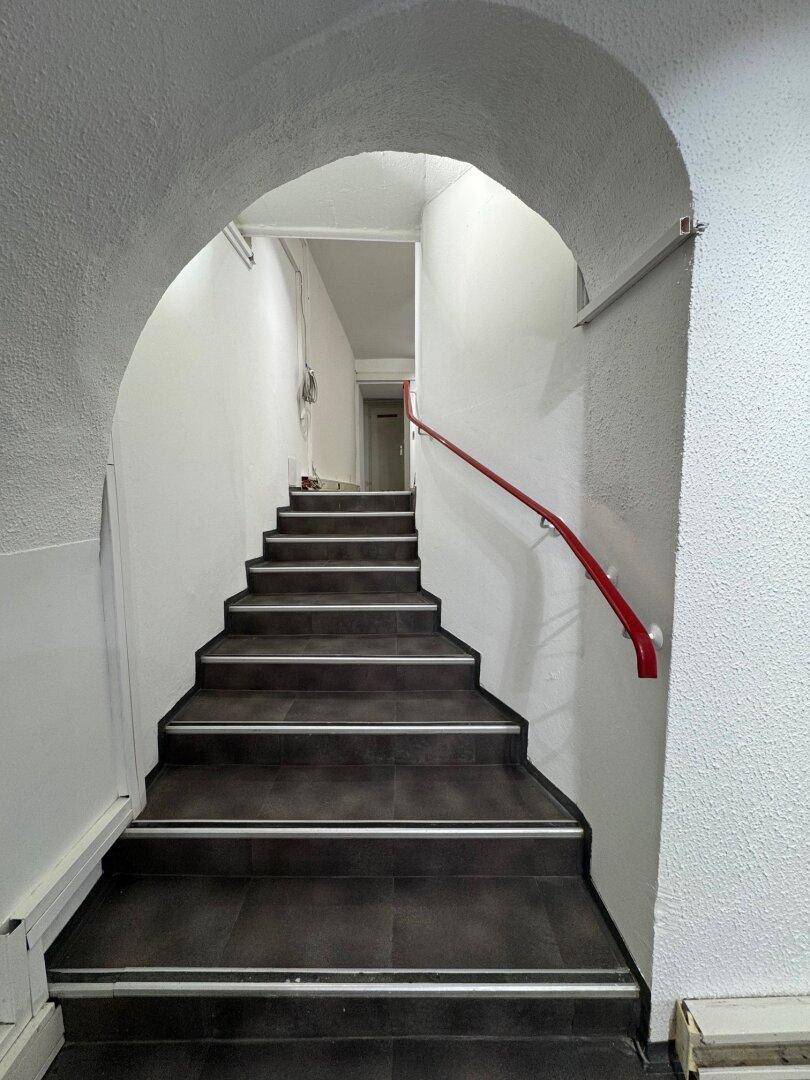 Dunkle Fliesentreppe mit rotem Handlauf unter einem Gewölbebogen, führt in die oberen Stockwerke.