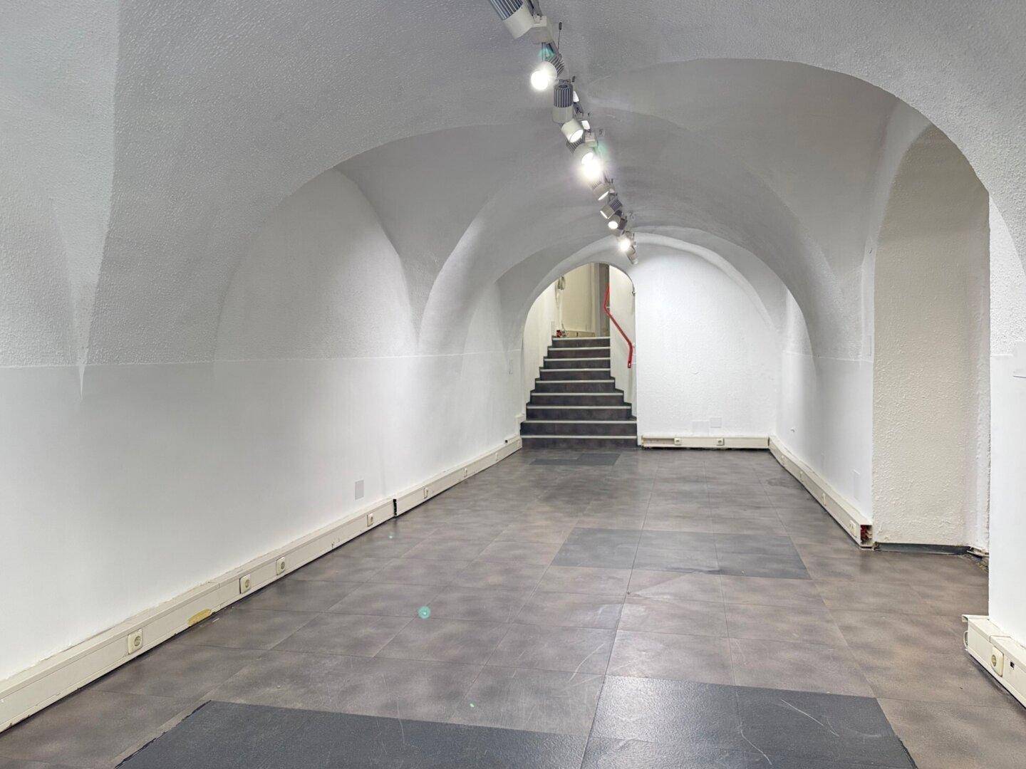 Langer Verkaufsraum mit Gewölbedecken, dunklem Fliesenboden und Treppenaufgang im Hintergrund.