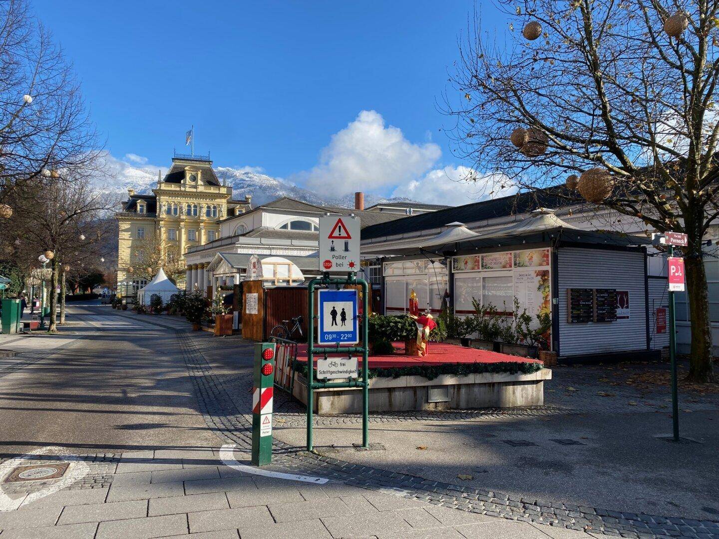Blick auf eine belebte Straße mit historischen Gebäuden und modernen Kiosken, umgeben von kahlen Bäumen und schneebedeckten Bergen im Hintergrund unter blauem Himmel.