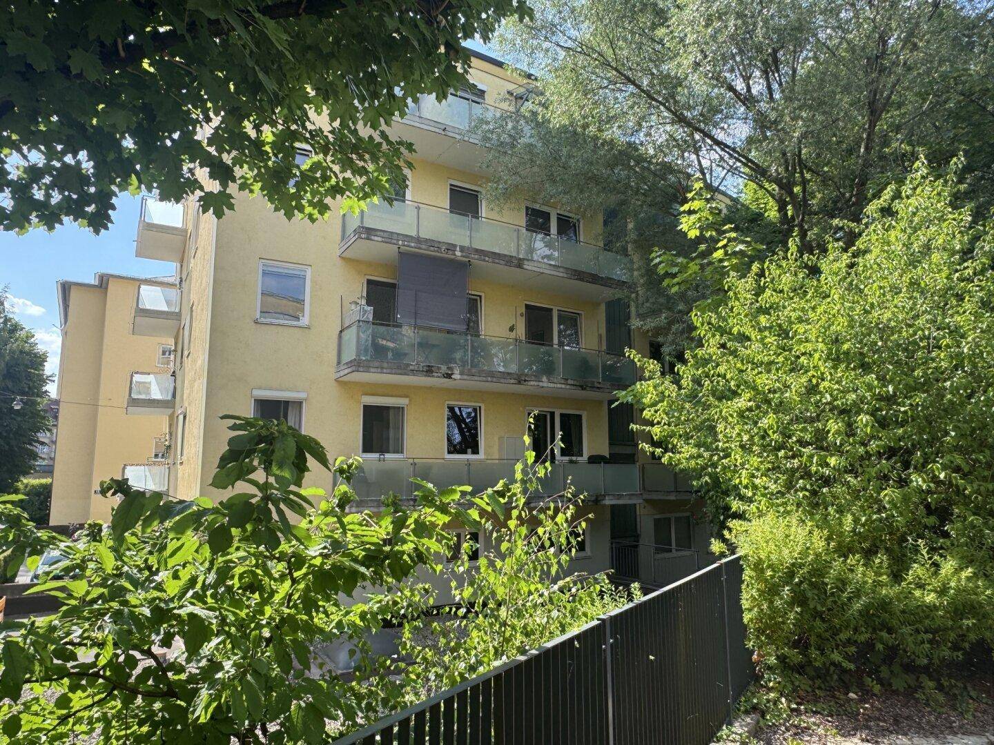 Mehrstöckiges Apartmentgebäude mit gelber Fassade und mehreren Balkonen, umgeben von üppigem Grün.