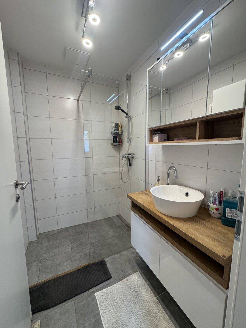 Modernes Badezimmer mit begehbarer Dusche, Waschtisch und Spiegelschrank, hell gefliest.