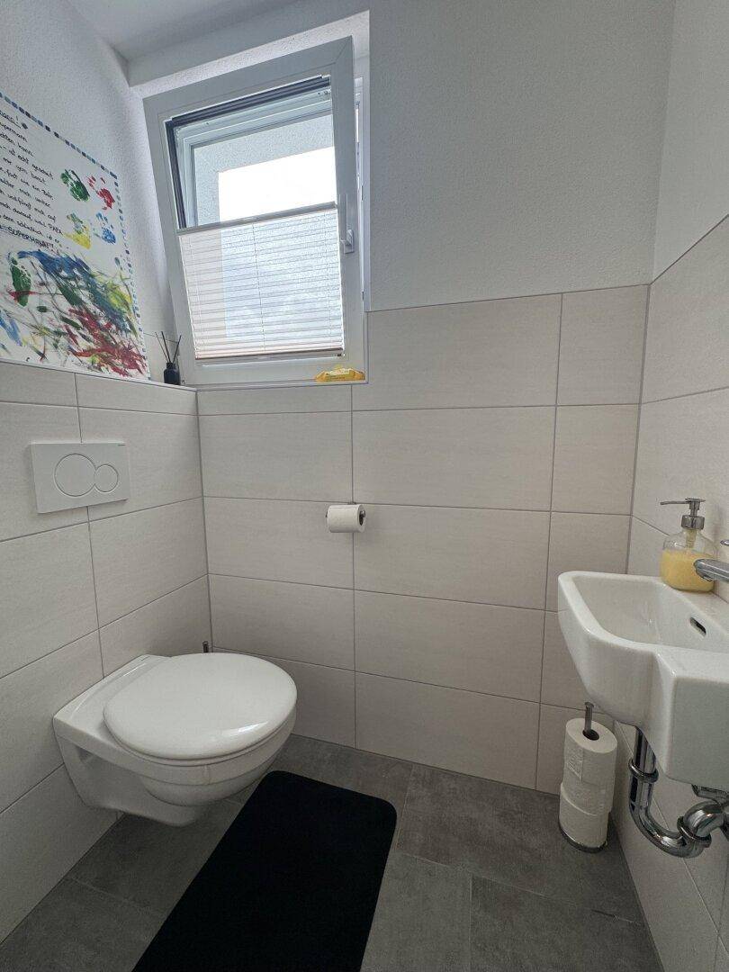 Helles Gäste-WC mit Wand-WC, kleinem Waschbecken und Fenster für natürliche Belüftung.