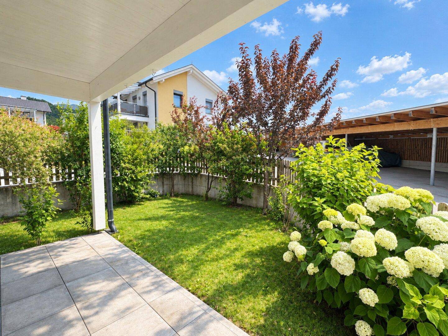Gepflegter Gartenbereich mit Rasenfläche, Sträuchern und einer überdachten Terrasse.