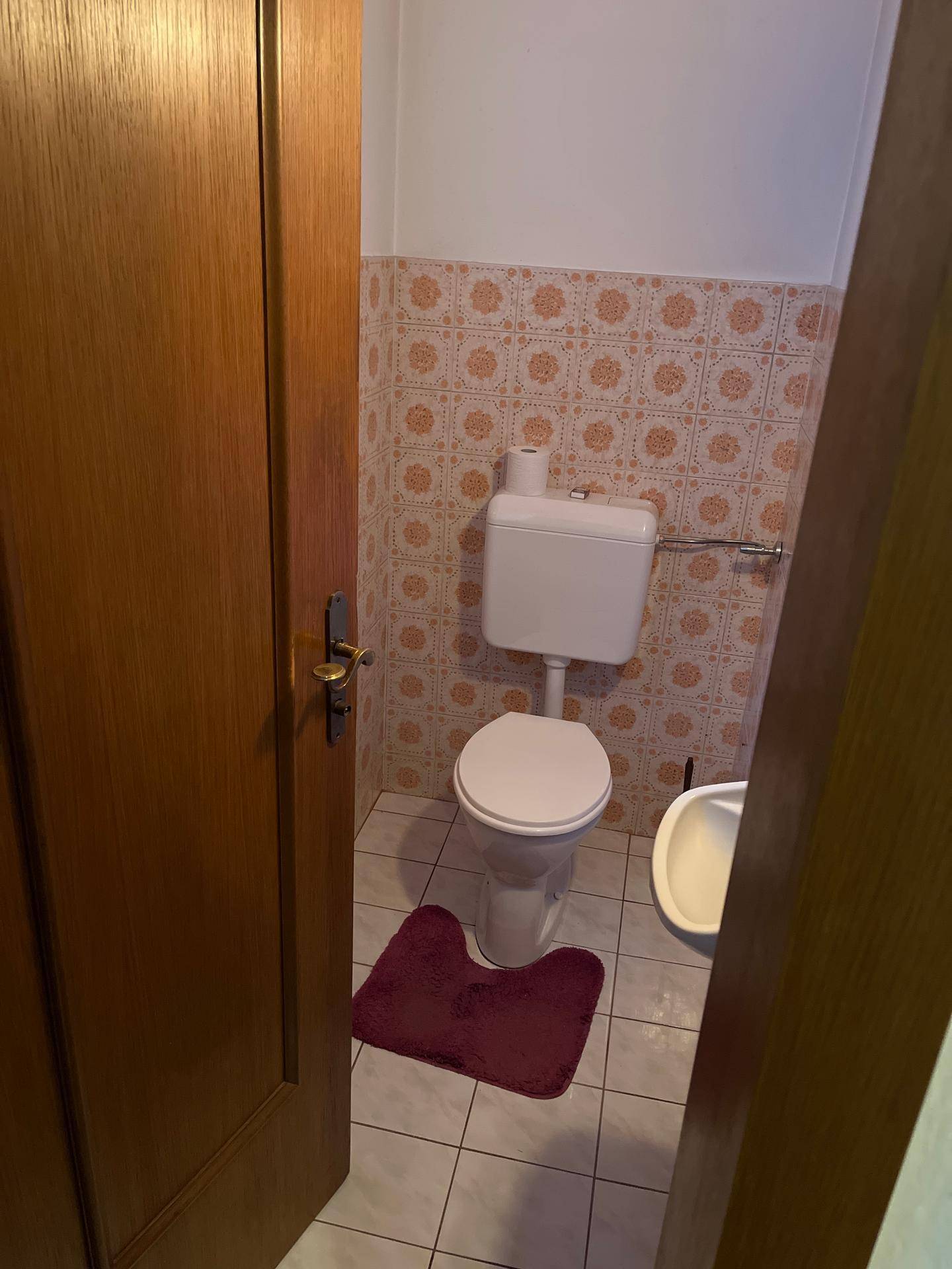 Kleines WC mit beigen Fliesen, Toilette und Bidet, durch eine Holztür zugänglich.