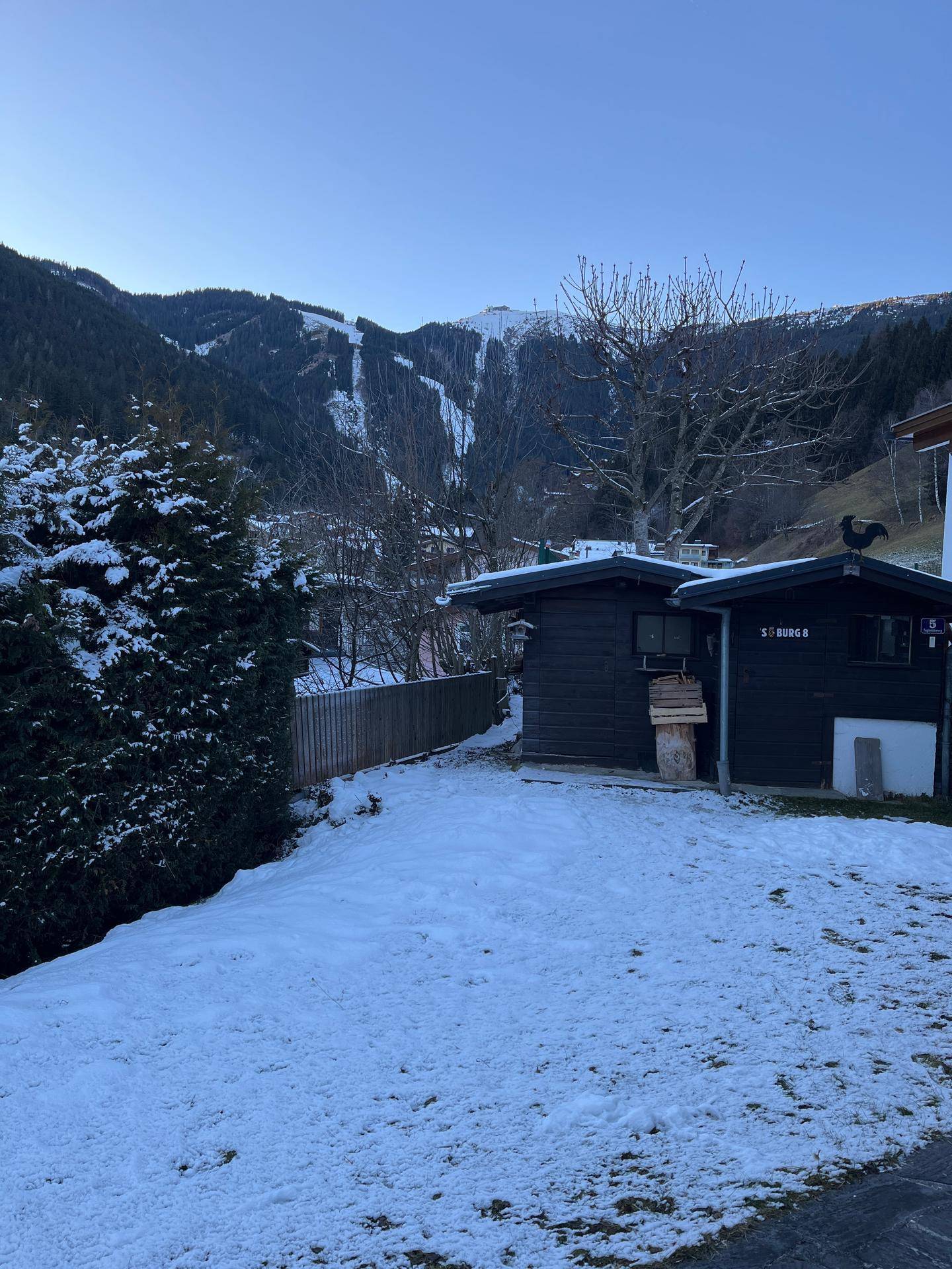 Verschneiter Gartenbereich mit zwei Holzschuppen und Blick auf die umliegende Berglandschaft.