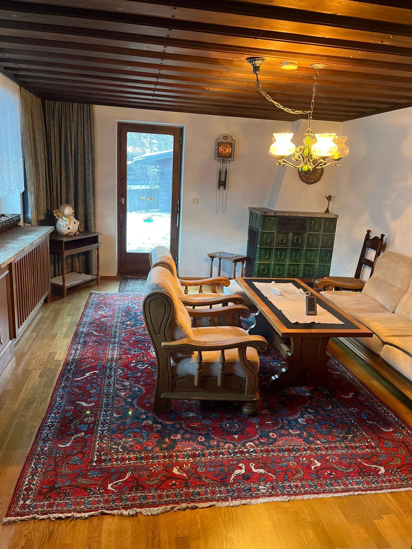 Geräumiges Wohnzimmer mit Kachelofen, Holzbalkendecke und traditionellem Teppichboden.