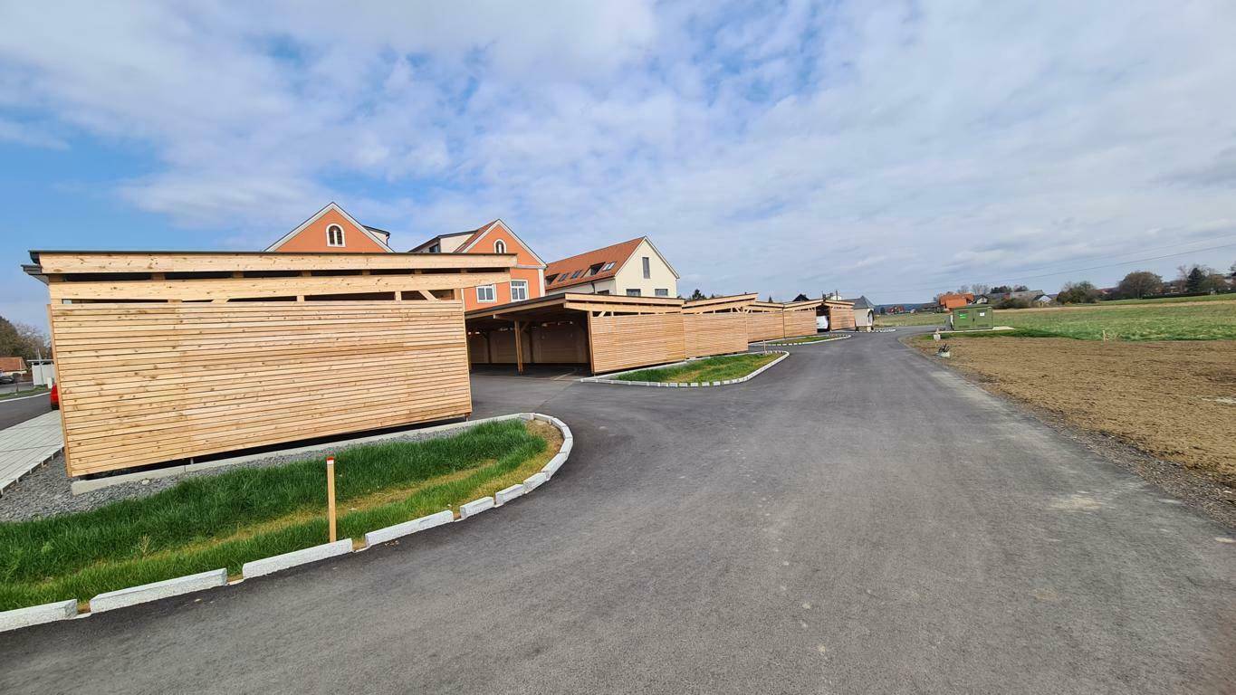 Lange Reihe von Carports aus Holz mit angrenzendem Feld und Wohngebäuden im Hintergrund.