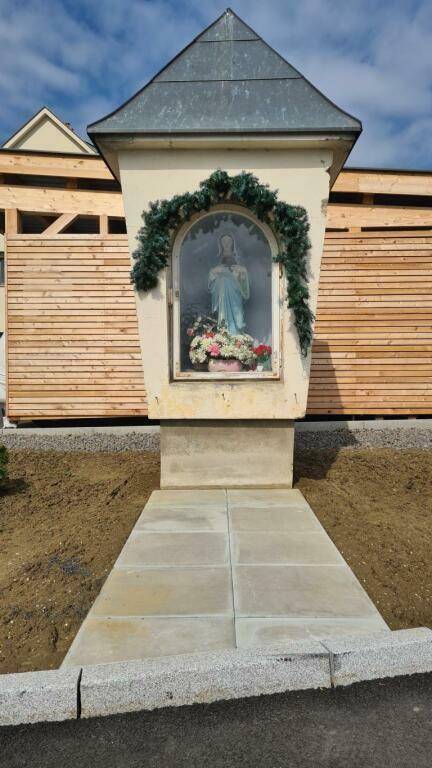 Religiöser Bildstock mit Marienstatue vor einem Gebäude mit Holzverkleidung.