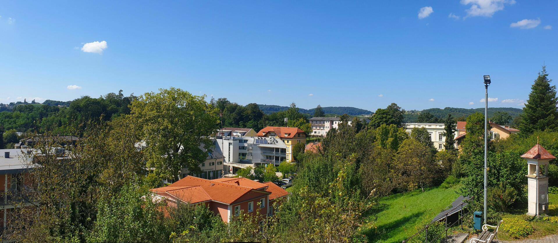Panoramablick über eine grüne Wohngegend mit Häusern und bewaldeten Hügeln unter blauem Himmel.