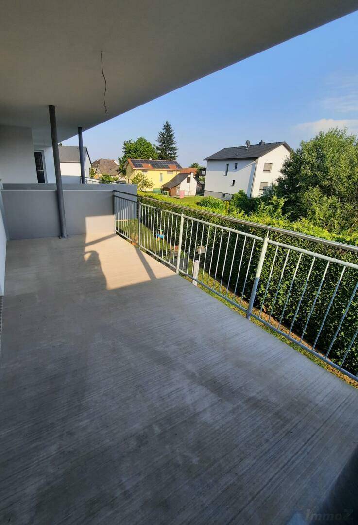 Geräumiger Balkon mit Betonboden, Metallgeländer und Blick auf grüne Hecken und Häuser.