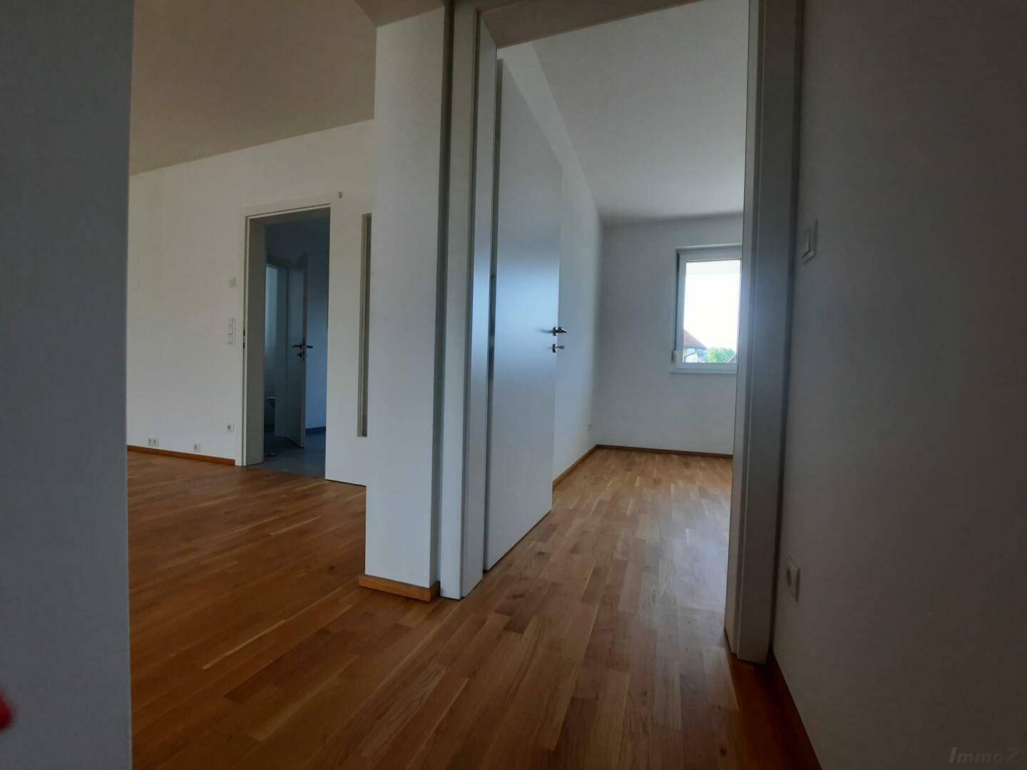 Leeres Zimmer mit hellem Echtholzparkettboden, weißen Wänden und einem Fenster mit Tageslicht.
