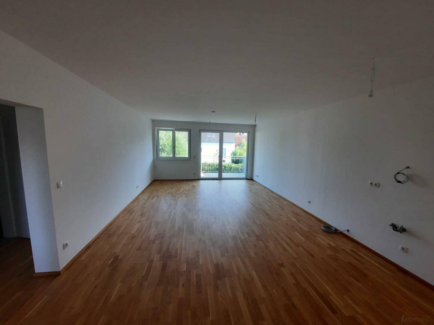 Großes, leeres Wohnzimmer mit Echtholzparkett, weißen Wänden und Zugang zu einem Außenbereich.