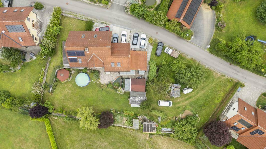 Luftaufnahme eines Einfamilienhauses mit großem Garten, oberirdischem Pool, Photovoltaikanlage auf dem Dach und mehreren Stellplätzen.