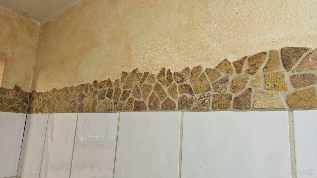 Detailansicht einer Kellerwand mit dekorativen Steinapplikationen über weißen Fliesen.