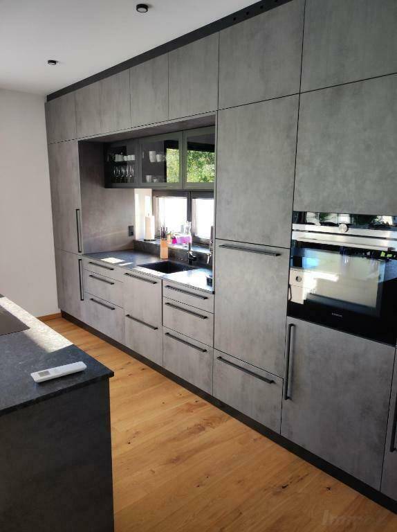 Moderne Einbauküche mit grauen Fronten, dunkler Arbeitsplatte und Holzboden, bietet natürlichen Lichteinfall und Blick ins Grüne.