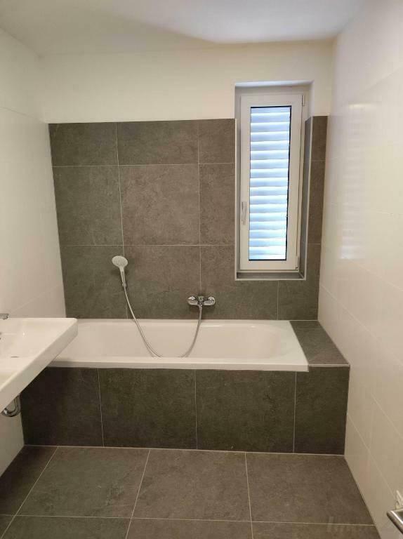 Modernes Badezimmer mit Badewanne, grauen Fliesen und Fenster für Tageslicht.