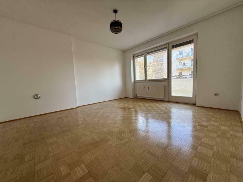 Leeres Wohnzimmer mit Parkettboden und Zugang zu einem kleinen Balkon.