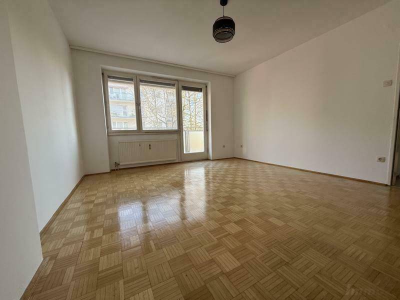 Geräumiges, leeres Wohnzimmer mit Parkettboden und viel Tageslicht durch große Fenster.