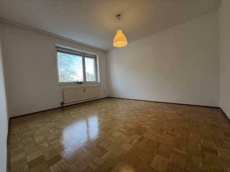 Helles, leeres Zimmer mit Parkettboden und einem Fenster mit Blick ins Grüne.