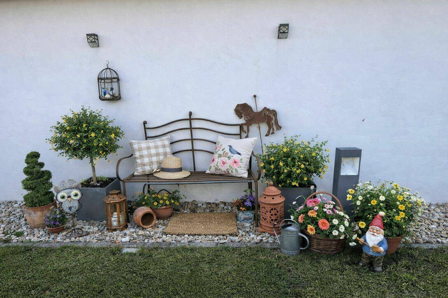 Gemütliche Gartendekoration mit einer Bank, Pflanzen und verschiedenen Figuren vor einer weißen Wand.