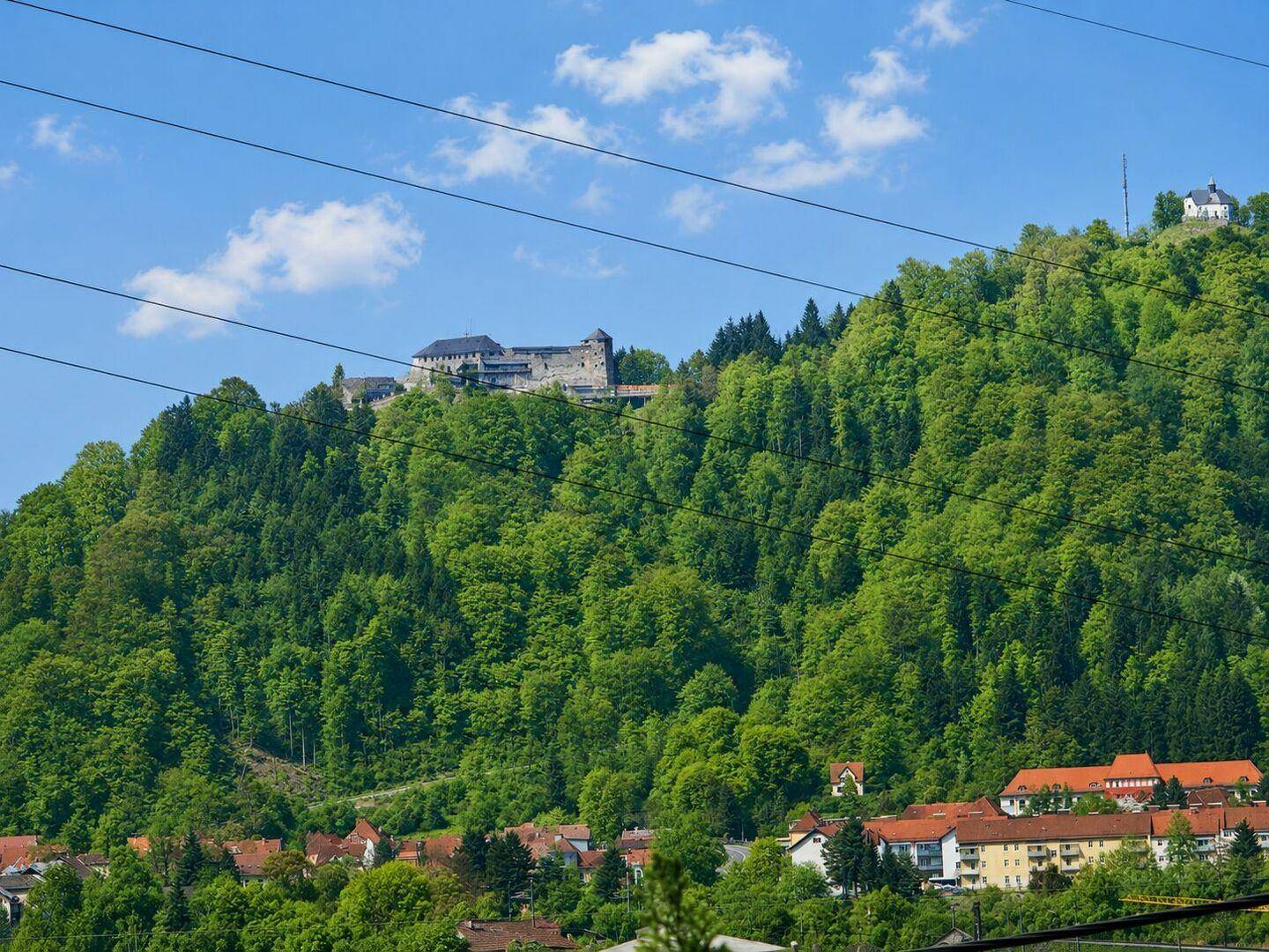 Historische Burg auf einem bewaldeten Hügel, über einer Siedlung unter blauem Himmel.