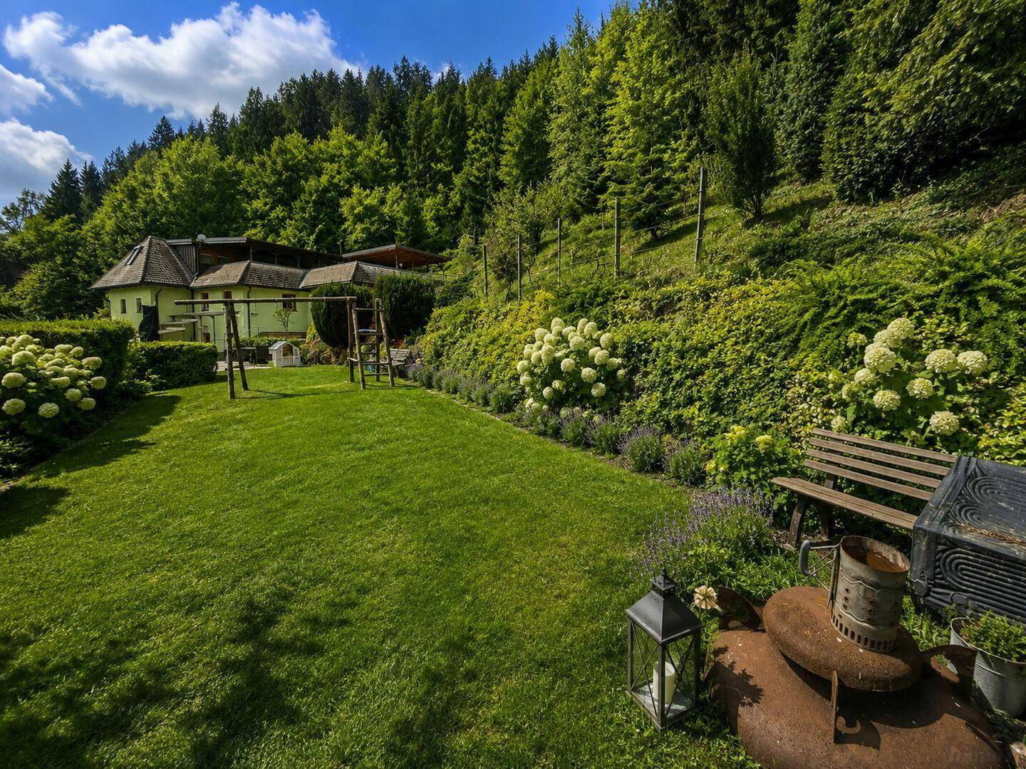 Gepflegter Garten mit Rasenfläche, Sitzbank und einem Grillbereich, umgeben von blühenden Pflanzen.