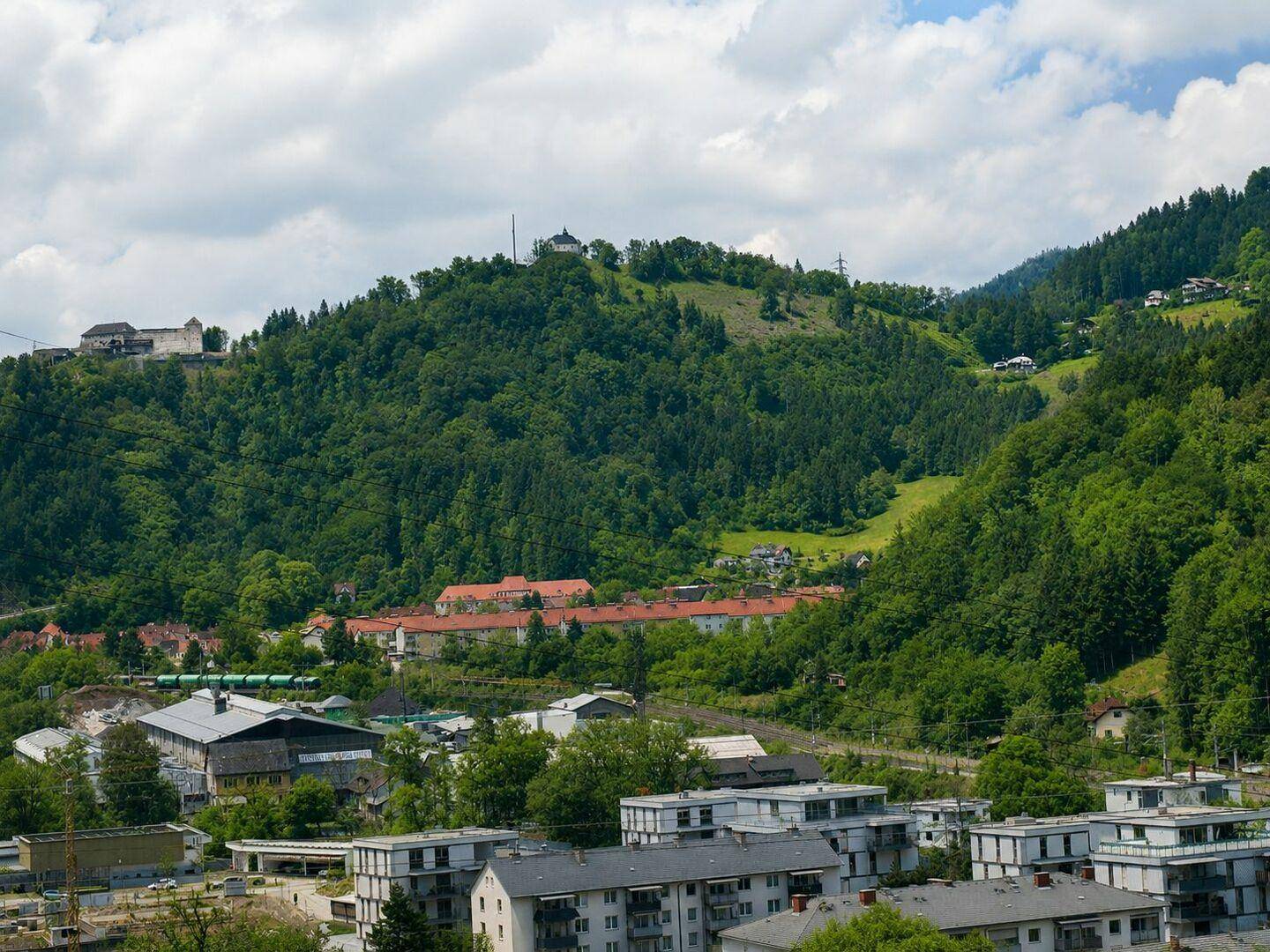 Panoramablick auf eine Stadt im Tal, umgeben von bewaldeten Hügeln und Bergen unter blauem Himmel.