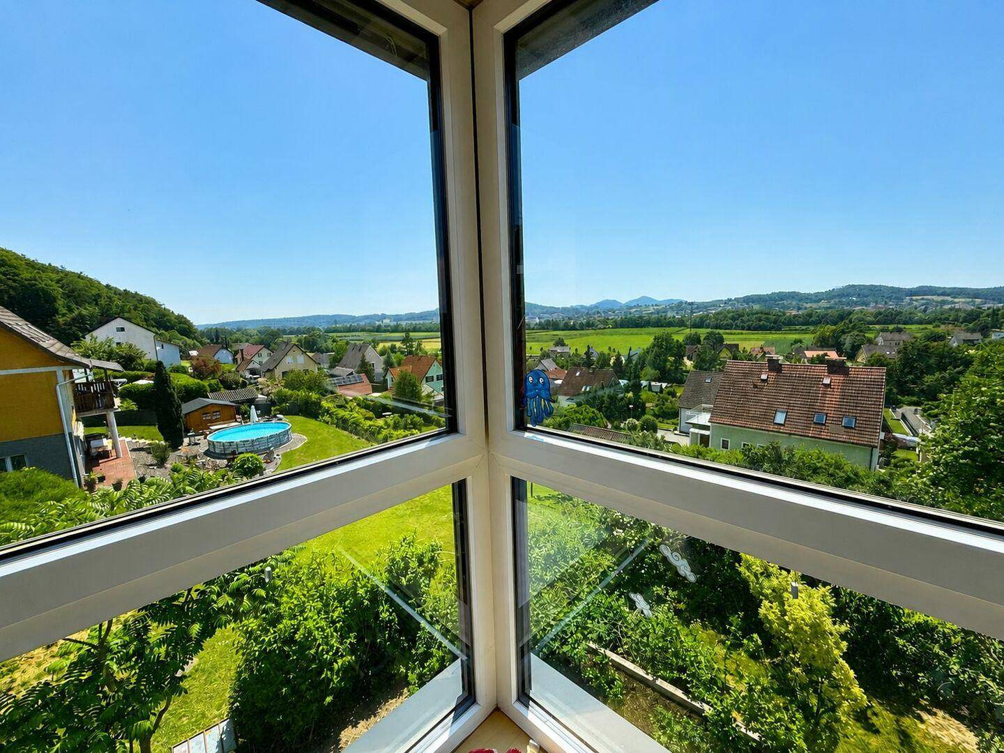 Panoramablick aus großen Fenstern auf die grüne Umgebung und die umliegenden Häuser.