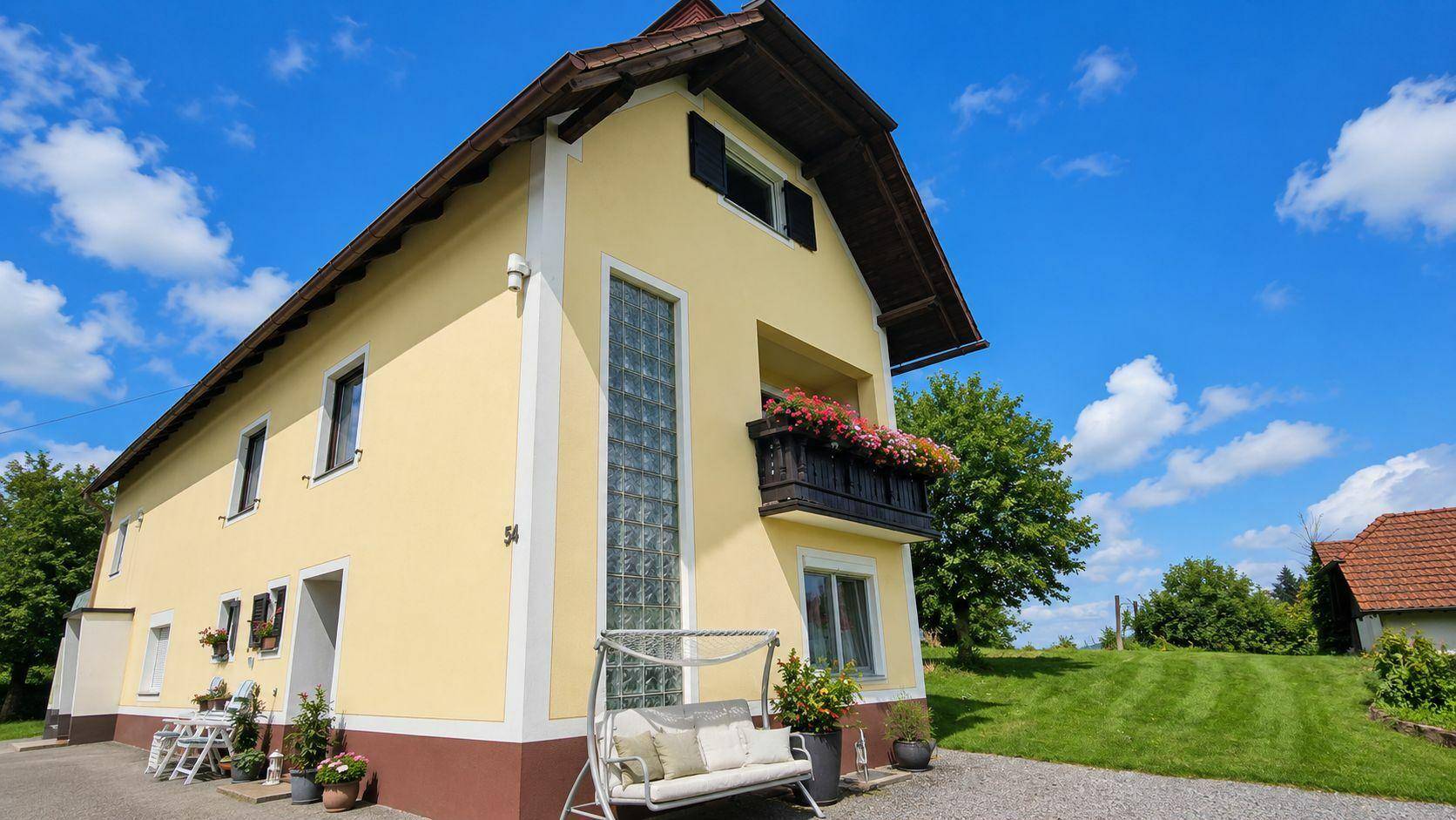 Gelbe Hausfassade mit Balkon und Schaukelbank im Vorgarten unter blauem Himmel.