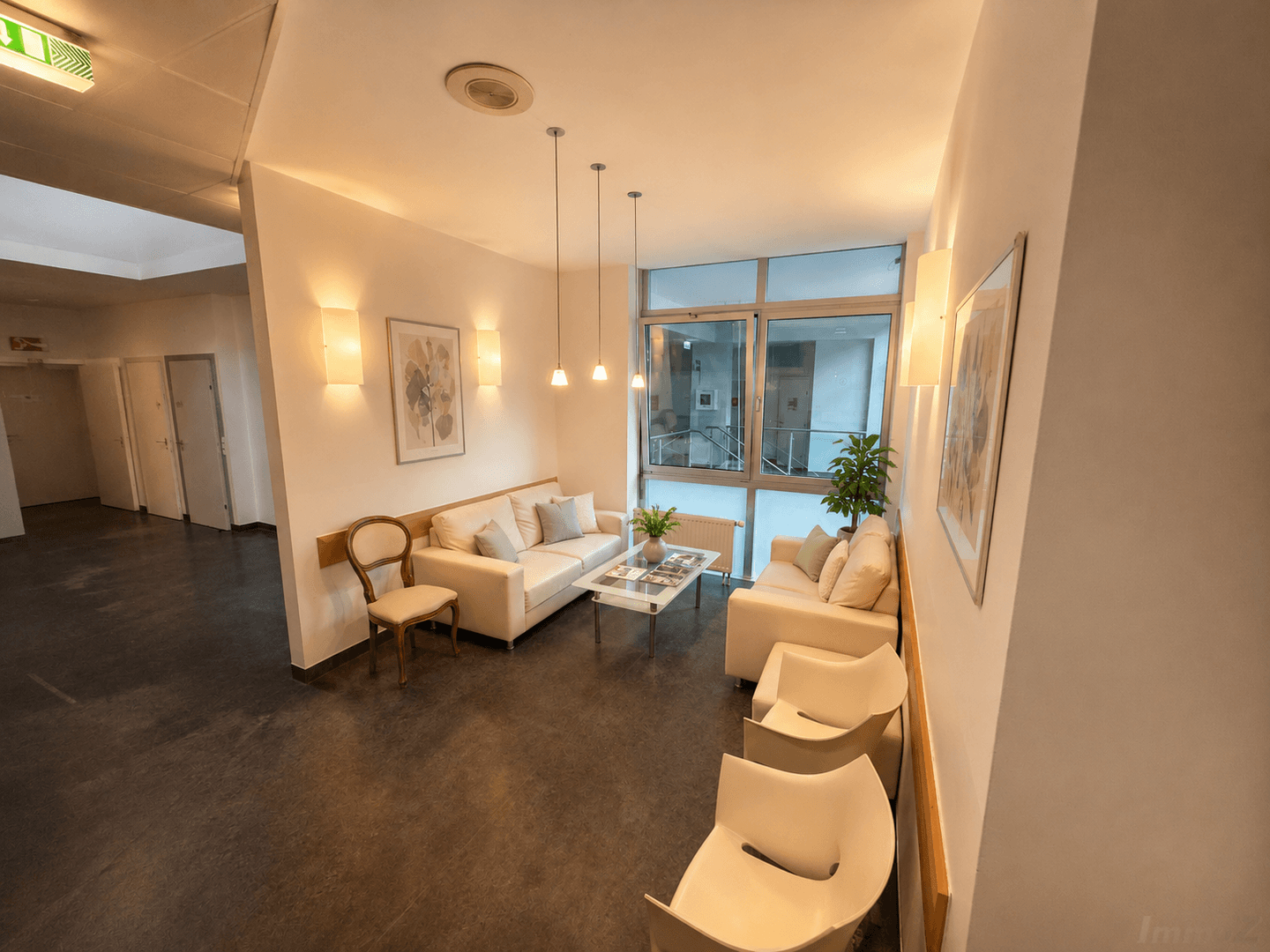 Komfortabler Wartebereich mit weißen Sofas, Glastisch und Blick auf das Treppenhaus durch große Fenster.