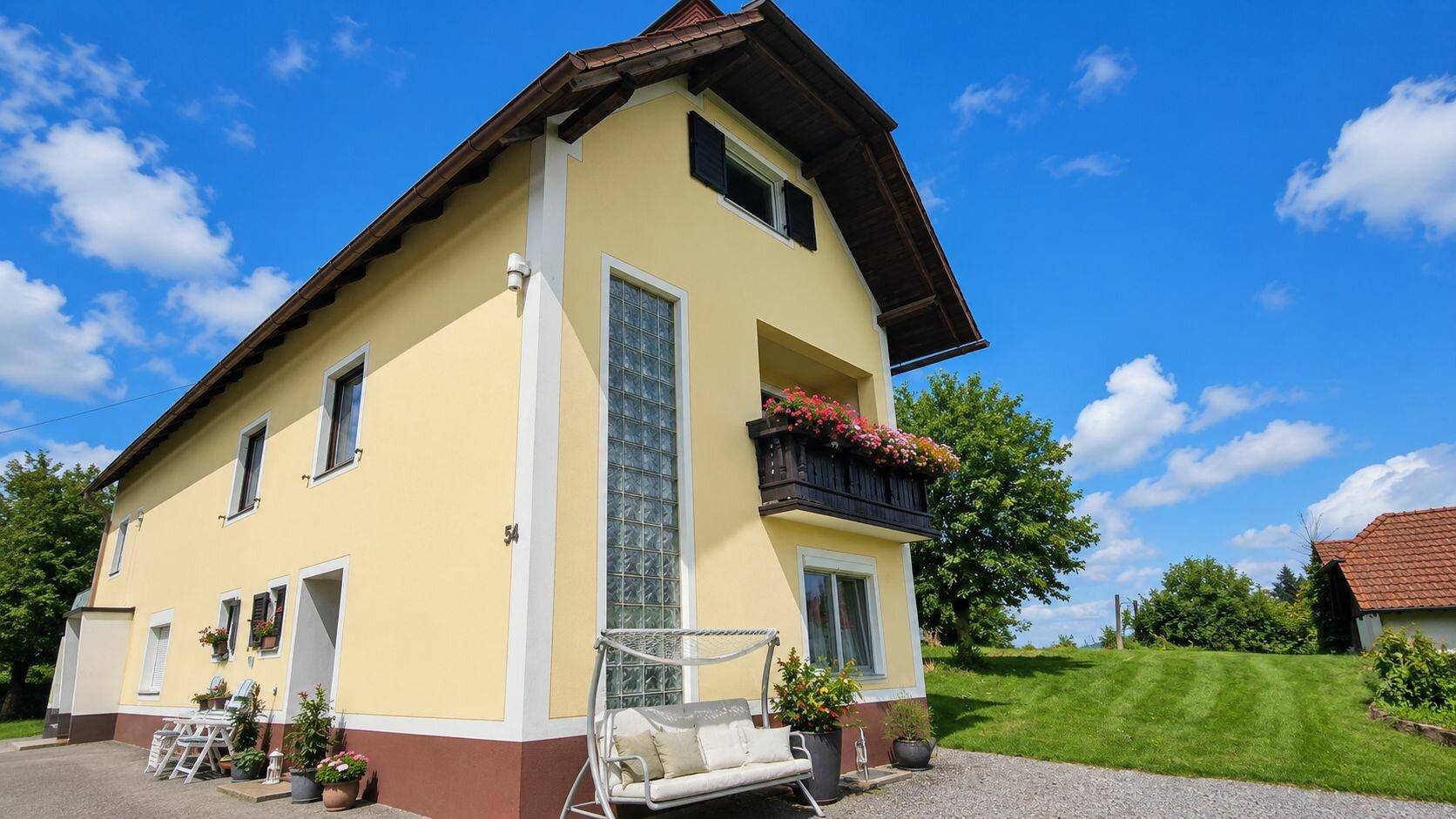 Ansicht des Hauses mit gelber Fassade, braunem Sockel und einem blumengeschmückten Balkon.