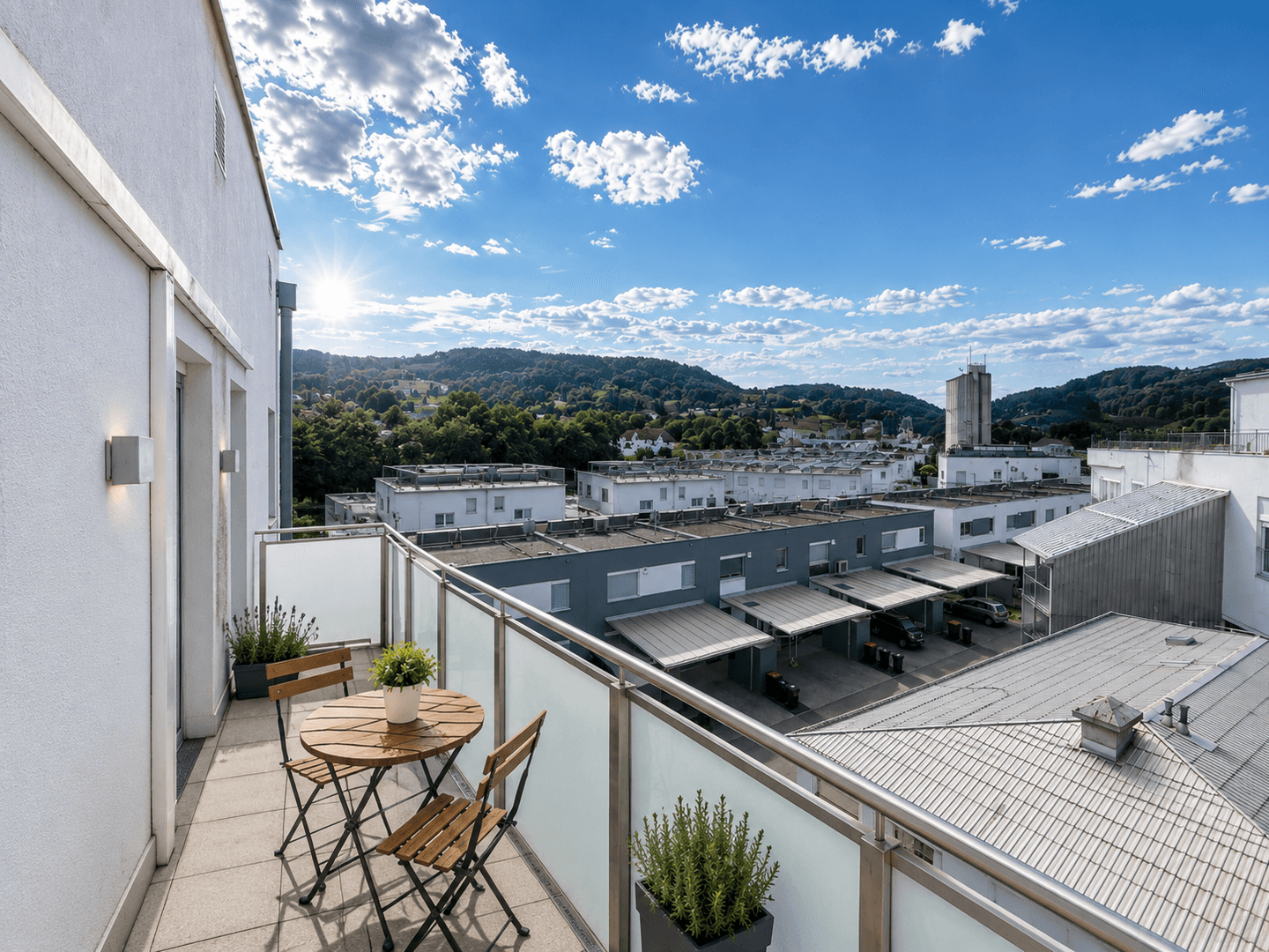 Sonniger Balkon mit kleinem Holztisch und Stühlen, bietet weiten Blick über die Umgebung und grüne Hügel.