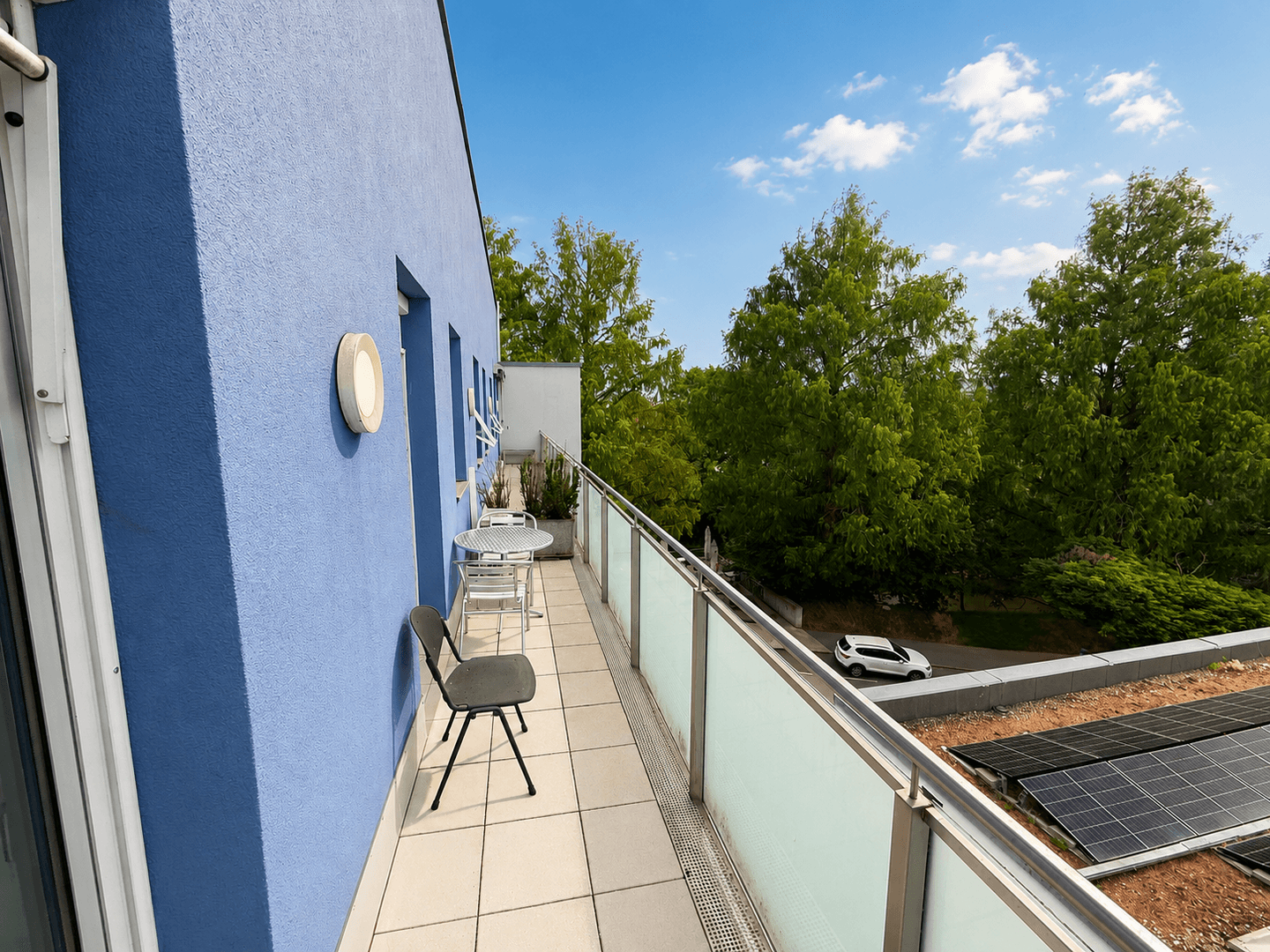 Langer Balkon mit blauer Fassade, Sitzgelegenheiten und Blick auf Solarpaneele auf dem Dach.