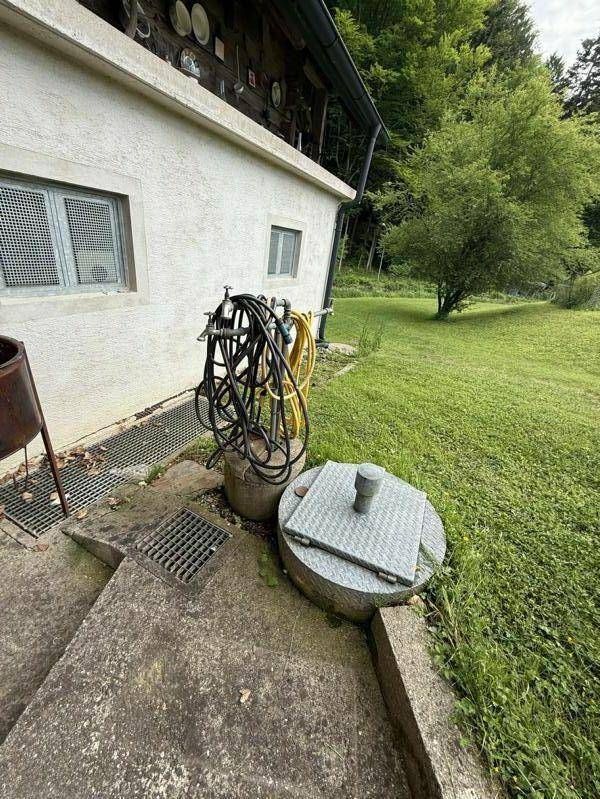 Seitenansicht des Hauses mit Wasseranschlüssen und Gartenschläuchen, umgeben von Grünflächen.