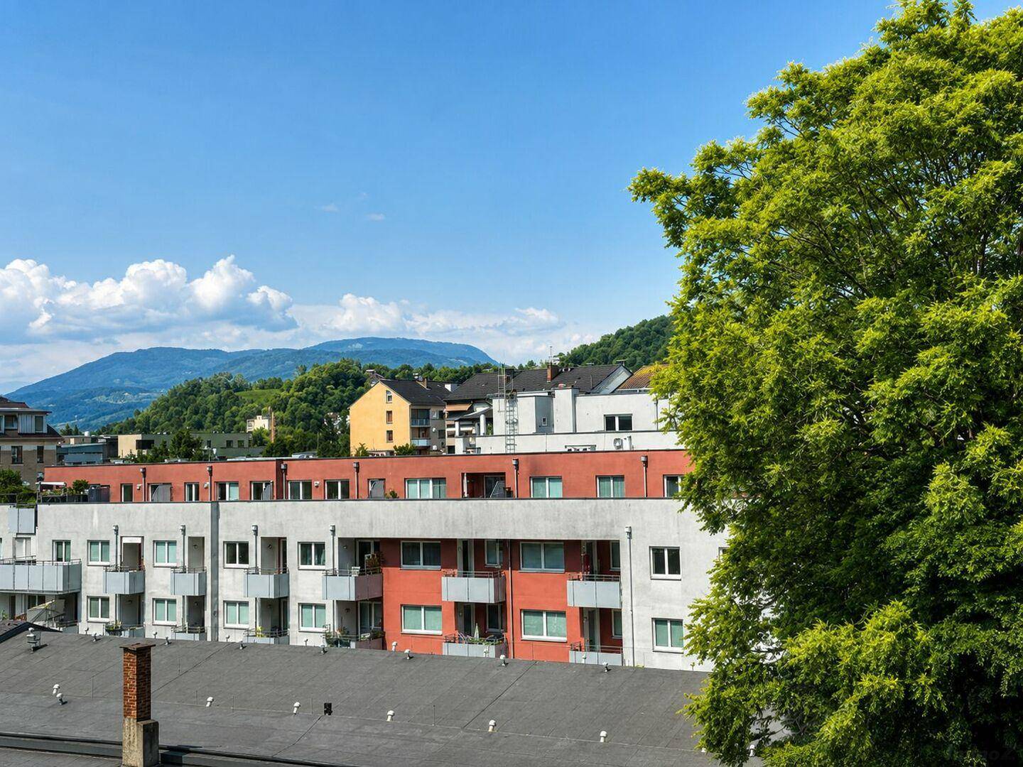 Panoramablick über Dächer und Wohngebäude mit grünen Hügeln und Bergen im Hintergrund.