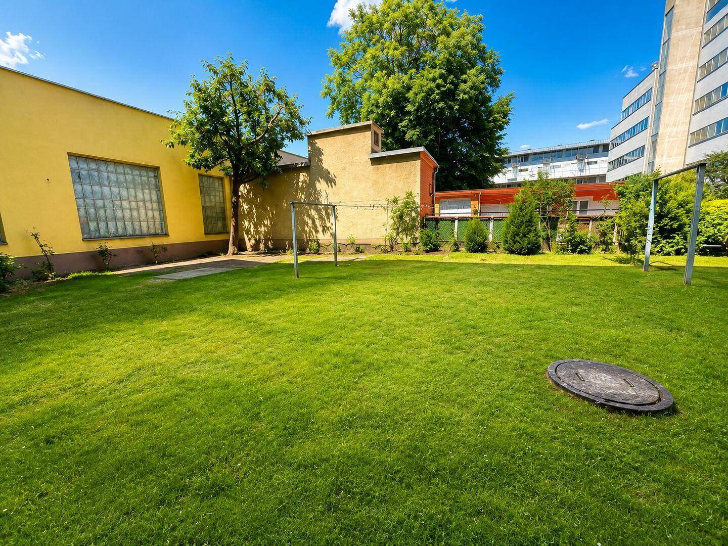 Weitläufige Rasenfläche im Gartenbereich, umgeben von Gebäuden unter blauem Himmel.