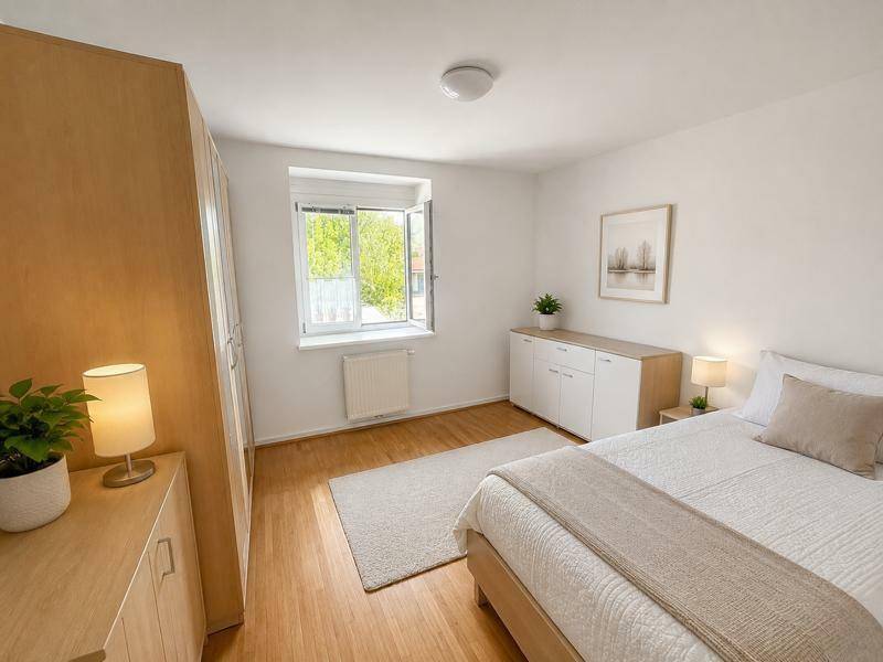 Helles Schlafzimmer mit Holzboden, weißer Einrichtung und großem Fenster mit Grünblick.