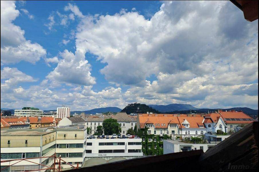 Weitläufiger Stadtblick über Dächer und Gebäude mit Bergen im Hintergrund unter blauem Himmel.